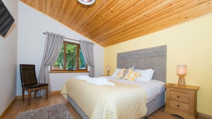 Ein Schlafzimmer mit Holzbalkendecke, einem Bett mit grauem Kopfteil, einem Nachttisch mit Lampe, einem Stuhl und einem Fenster mit Vorhängen.