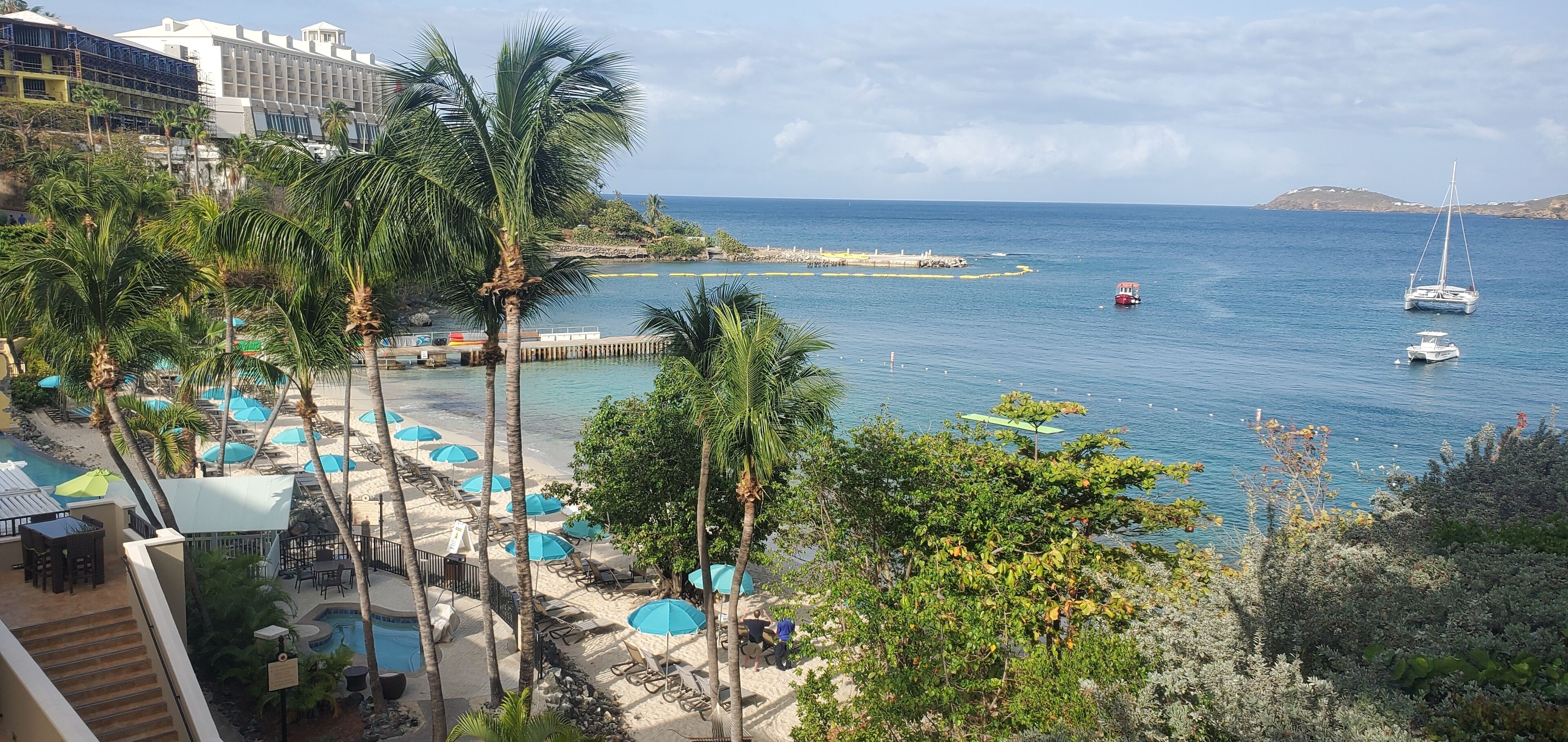 A beachside view with a pool area, palm trees, and a view of the sea with boats.