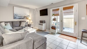 Una sala de estar con un sofá gris, una mesa de centro blanca, un televisor de pantalla plana montado en la pared y una estantería con diversos objetos.