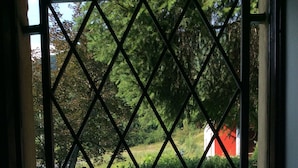 Vista per una finestra amb una reixa de dibuixos de diamant, que dona a una piscina i una zona de jardí amb un edifici vermell al fons.