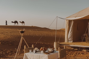 Un campement dans le désert, avec une tente, une table dressée avec de la nourriture, et des chameaux en arrière-plan.