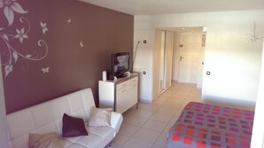 A hotel room with a white sofa, a TV on a stand, a bed with a patterned bedspread, and a door leading to a bathroom. - Comfortable studio apartment in Le Gosier (Le Gosier)
