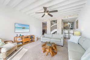 A living room with a sofa, a glass coffee table, a wooden side table, a television, and a staircase leading to a kitchen. - Cayman Wind and Sea! (West Bay)