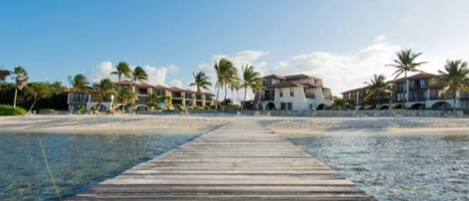 A wooden pier leading to a beachfront property with multiple buildings, palm trees, and clear skies.