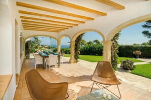Un patio al aire libre con una mesa y sillas, una zona cubierta con vigas de madera y vista al mar.