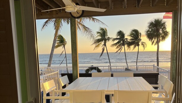 A deck with white furniture overlooking a beach with palm trees and a sunset. - Steps away from the sugar white sands and blue water!! (Fort Myers Beach)