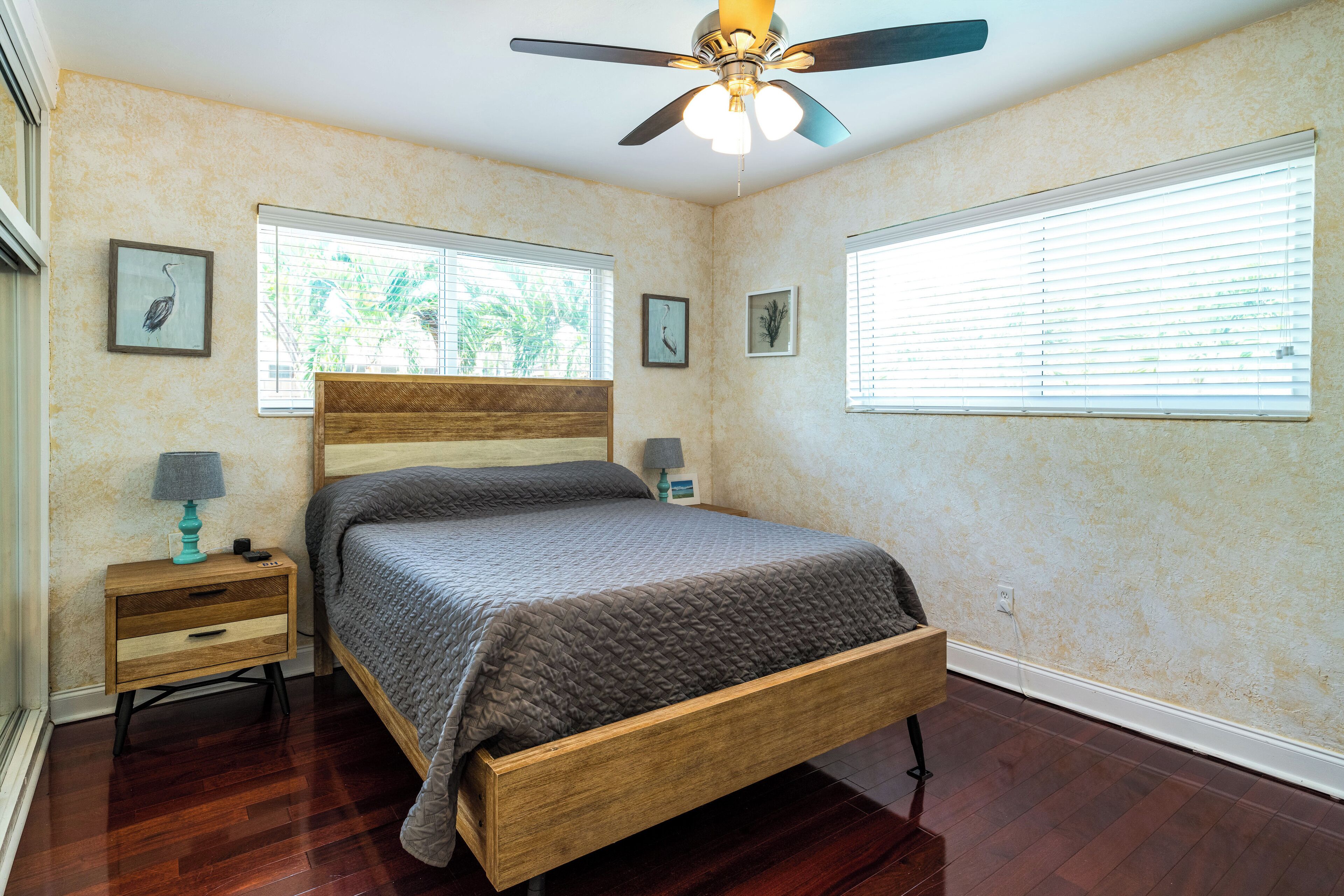 A bedroom with a wooden bed, two bedside tables, a ceiling fan, and two windows with blinds.