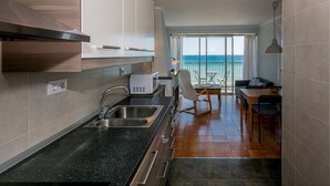 Eine kompakte Küche mit Spüle, Mikrowelle und Holzschränken. Ein Essbereich mit Tisch und Stühlen ist zu sehen, der auf einen Balkon mit Meerblick führt.