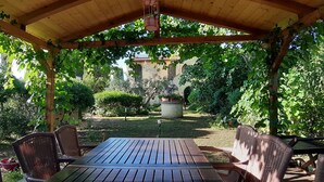 Un porxo cobert amb una taula de fusta i cadires de vimetla, envoltat de vegetació exuberant i amb vista a un edifici al fons.