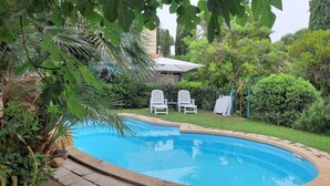 Una piscina envoltada de vegetació exuberant, amb dues cadires blanques i una taula coberta al fons.