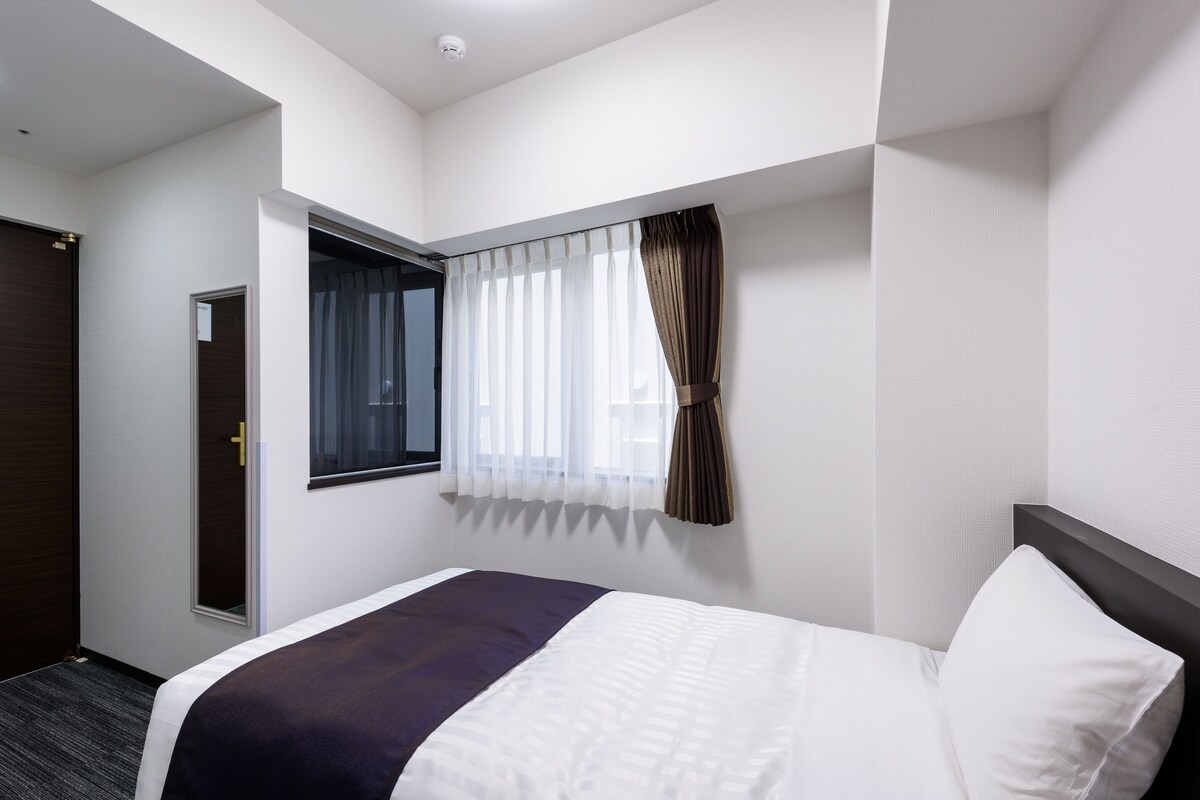 A hotel room with a bed, a window with curtains, a door, and a wall-mounted light fixture.