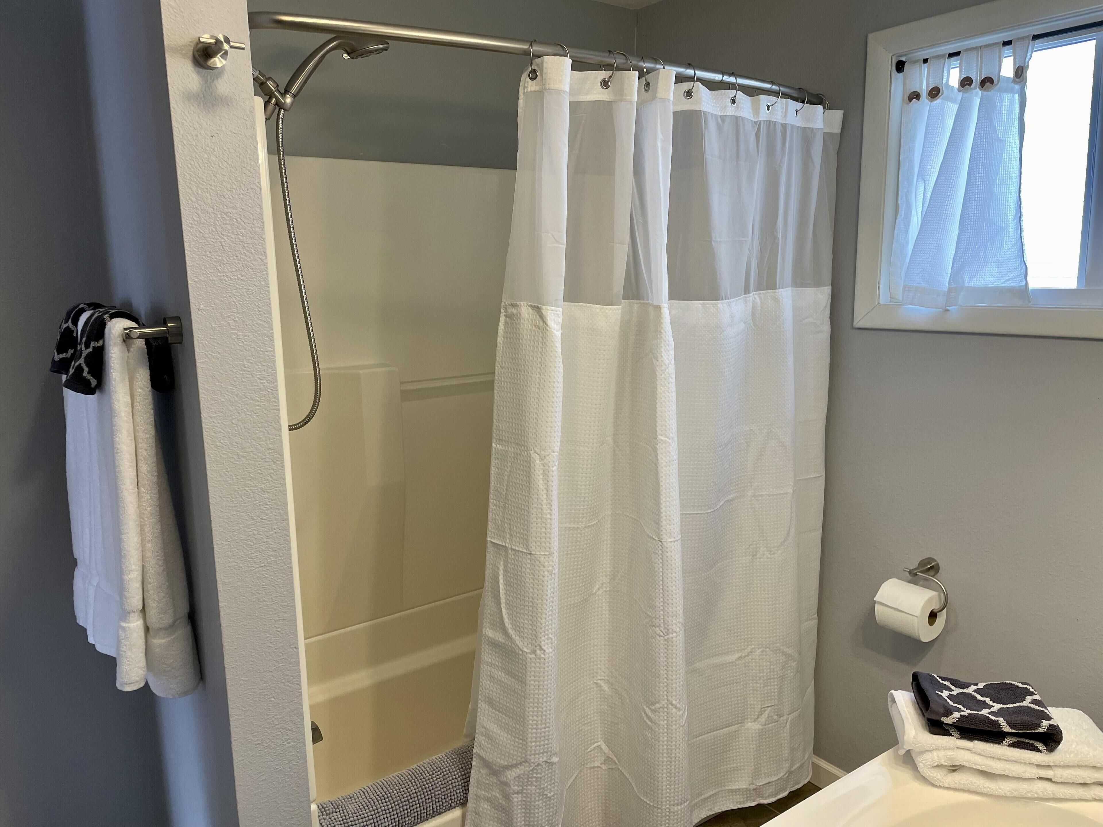 Un baño con cortina de ducha, toallero, dispensador de papel higiénico y una ventana con cortinas.