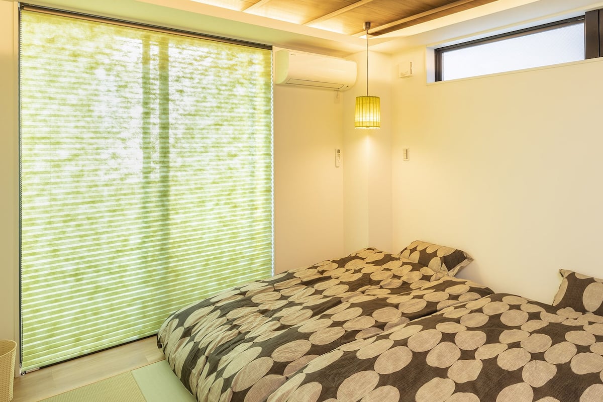 A bedroom with a bed, a window with blinds, a wall-mounted light, and a small basket.