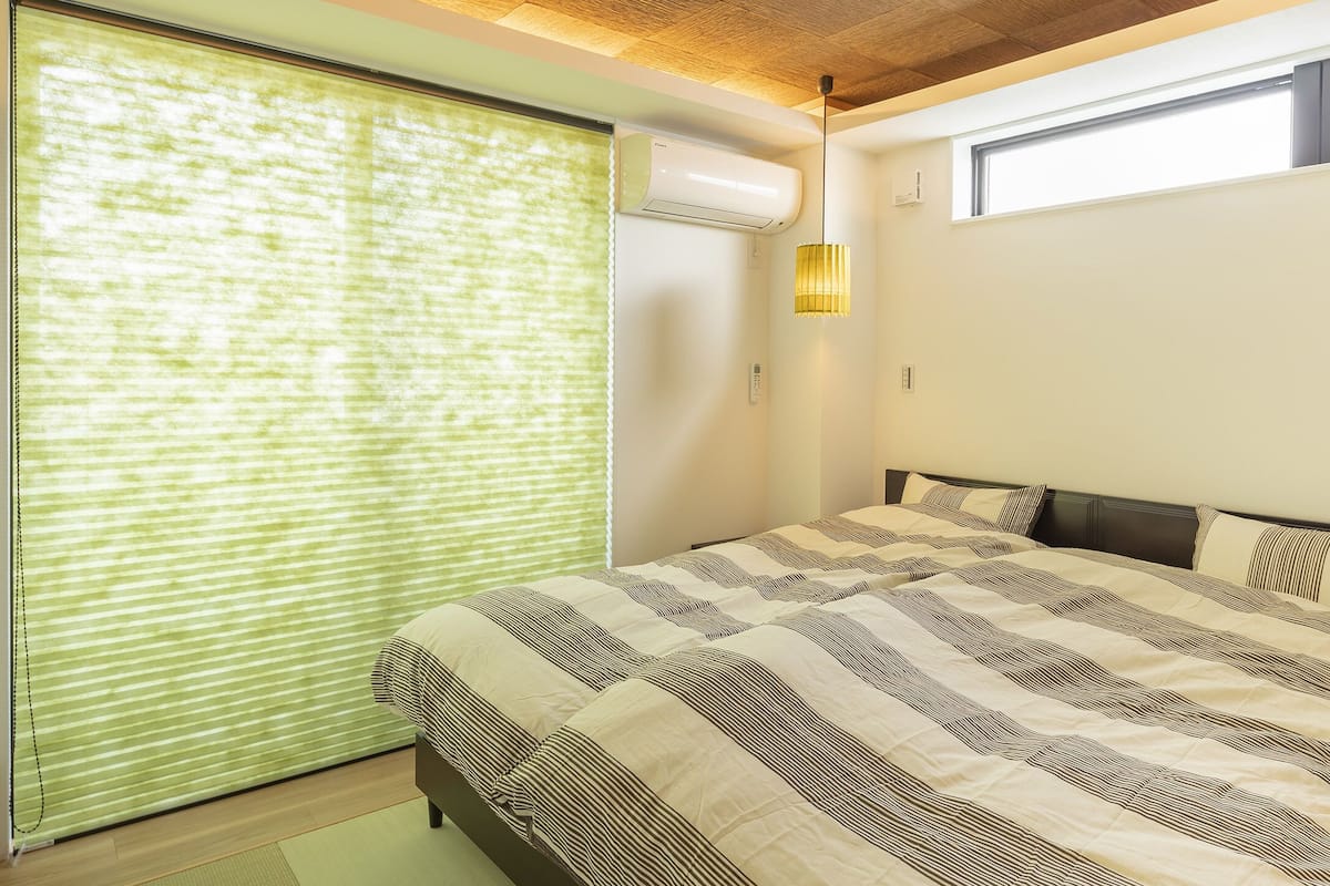 A bedroom with a large bed, a window with blinds, an air conditioning unit, and a small window above the bed.