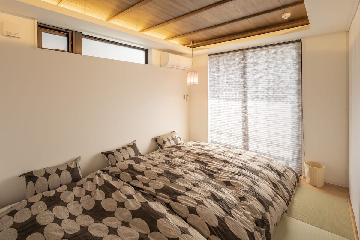 A bedroom with a large bed, a window with blinds, a ceiling light, and a small basket by the window.