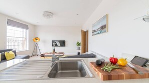 A modern living room with a built-in kitchen area, a flat-screen TV, and a dining table with a cutting board and vegetables. - South Quay Penthouse - 2 Bedroom - Parking - River (Great Yarmouth)