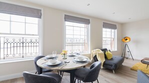 A dining area with a round table set for four, a sofa, a floor lamp, and large windows with metal railings. - South Quay Penthouse - 2 Bedroom - Parking - River (Great Yarmouth)