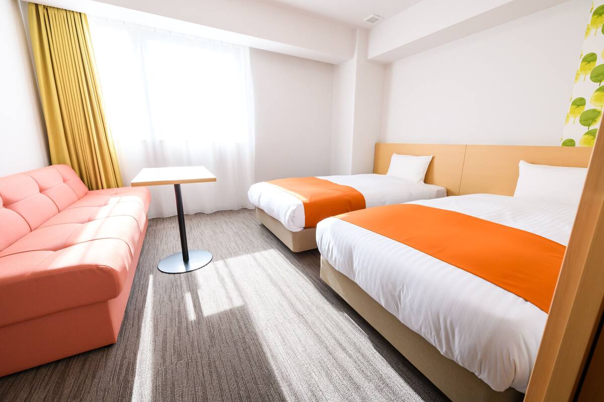 Ein Hotelzimmer mit einem rosa Sofa, zwei Betten mit orangefarbener Bettwäsche, einem kleinen Tisch und einem Fenster mit Vorhängen.