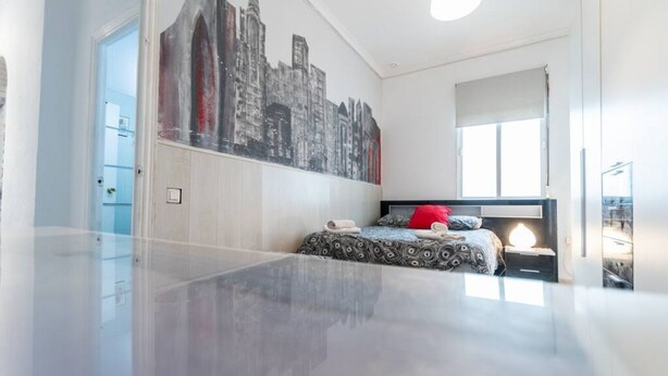 A modern bedroom with a cityscape mural, a bed with patterned bedding, a bedside lamp, and a large window. - Apartamento La Perla flamenca (Cádiz)