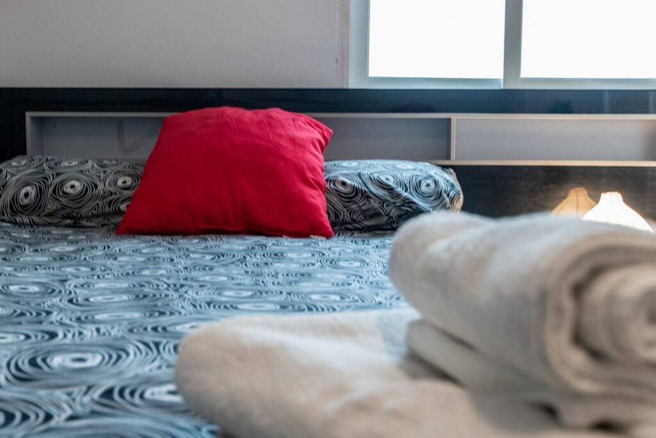 A bed with a patterned bedspread, a red pillow, and a rolled-up towel.