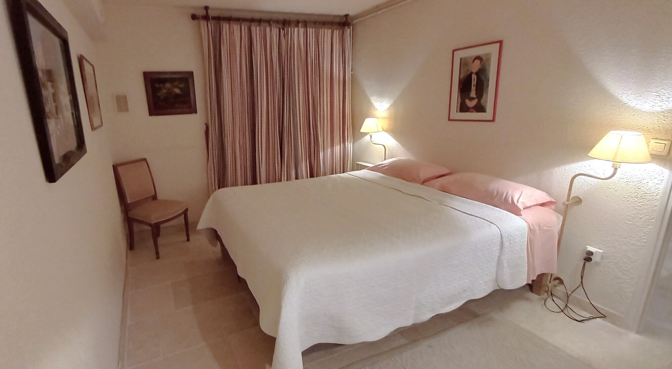 A bedroom with a bed, two bedside lamps, a chair, a window with curtains, and two framed pictures on the wall.