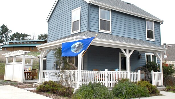Een tweelaags huis met een blauwe gevel, een witte veranda en een blauwe vlag met een wit embleem.