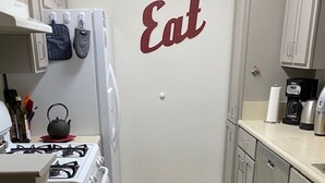 Een kleine keuken met een gasfornuis, een waterkoker en een koelkast. Een rood-wit gestreept vloerkleed ligt op de vloer, en op de muur staat het woord 'Eat'.