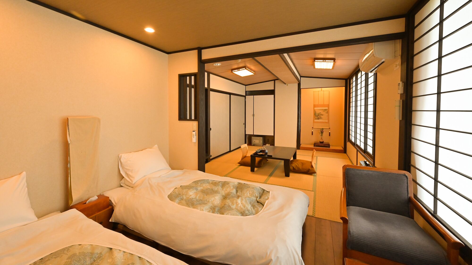 A traditional Japanese-style hotel room with tatami flooring, a futon bed, a desk with a chair, and sliding doors.