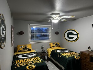 Ein Schlafzimmer mit zwei Betten, einem Deckenventilator, einem Fenster mit Jalousien und einer Wand mit großem Packers-Logo.