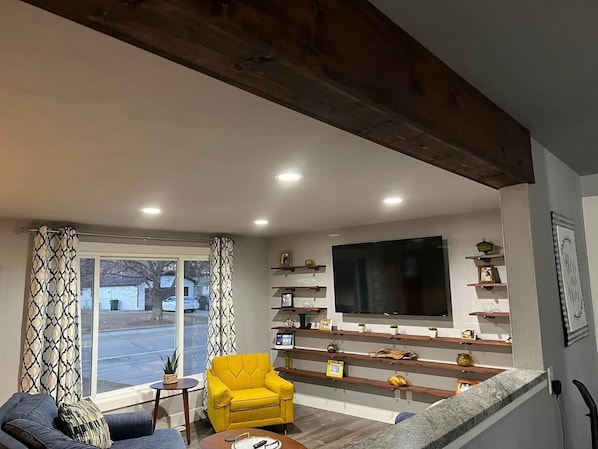 Una sala de estar con un sofá, un sillón amarillo, un televisor y estantes con objetos decorativos.