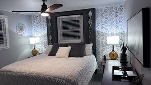Une chambre à coucher comprenant un lit, deux tables de chevet, un ventilateur de plafond et une fenêtre avec des stores.
