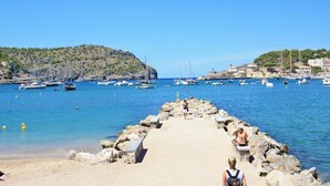 Escena costera con un muelle rocoso que lleva a aguas tranquilas, barcos y una colina con edificios al fondo.
