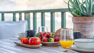 Ein Tisch mit Frühstücksangeboten, darunter Tomaten, Erdbeeren, Brot und ein Glas Orangensaft, mit einem Blumentopf und einem schönen Ausblick im Hintergrund.