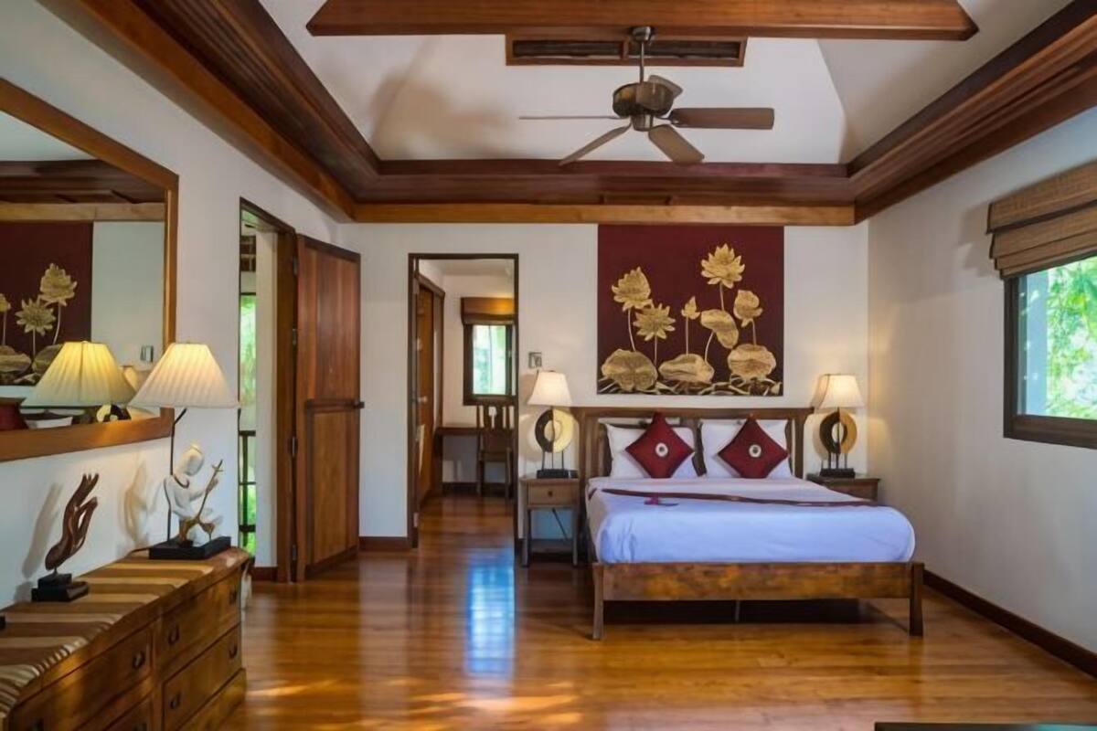 A bedroom with a bed, a ceiling fan, a wooden dresser, and a wall with a floral painting.