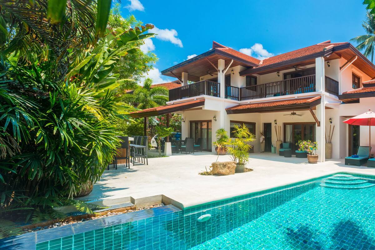A two-story house with a swimming pool, outdoor seating, and tropical plants.