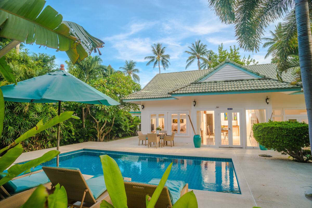 A pool area with lounge chairs, a dining table, and a house with a tiled roof.