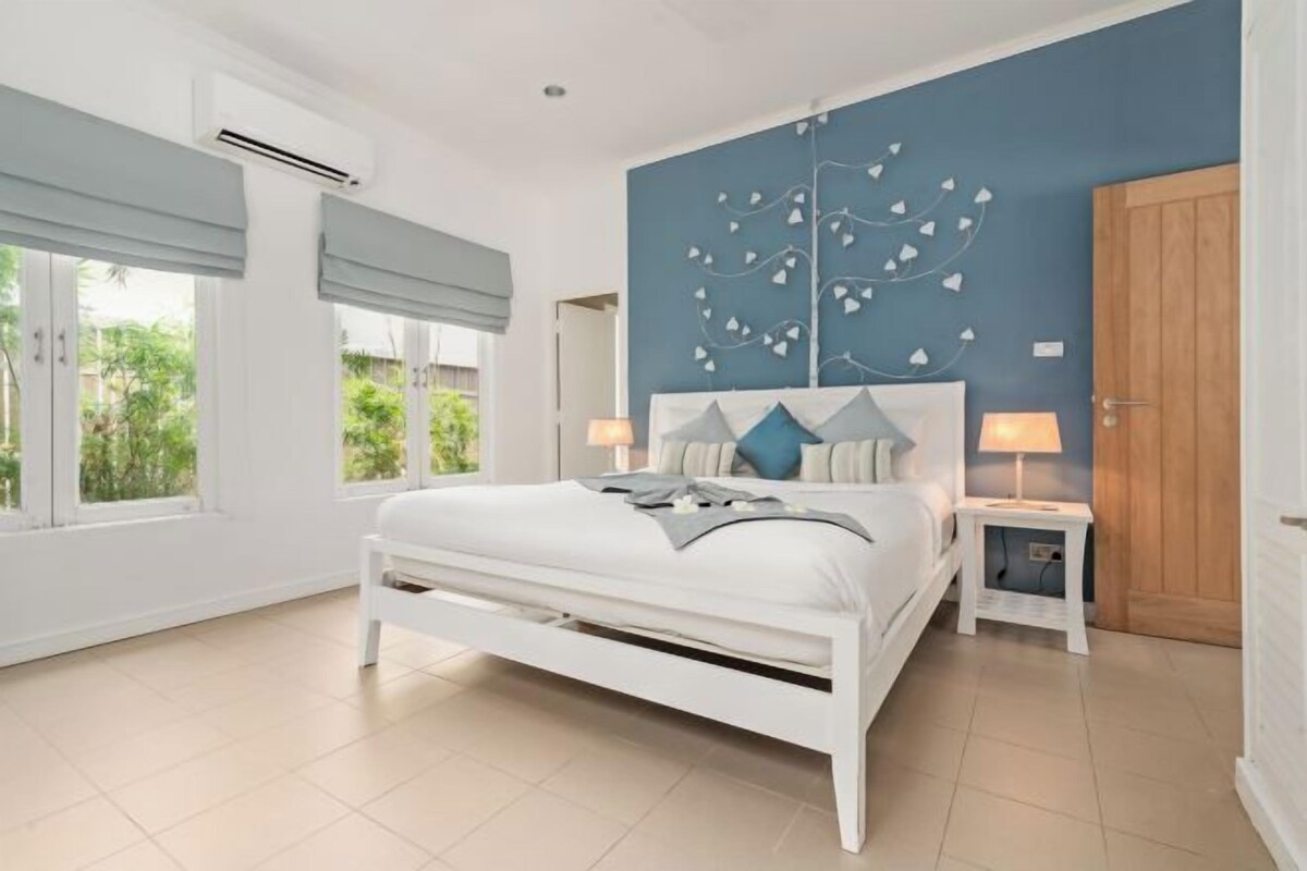 A bedroom with a large bed, a wall-mounted air conditioner, a bedside table with a lamp, and a view of greenery outside.
