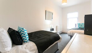 A hotel room with a bed, bedside table, lamp, and a window. - Furnished 2BR House in Leigh | Free Parking & Fast WiFi | Long Stays Welcome (Leigh)