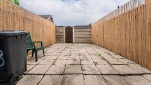 A courtyard with a stone floor, a wooden fence, a bench, and a trash bin. - Furnished 2BR House in Leigh | Free Parking & Fast WiFi | Long Stays Welcome (Leigh)