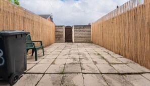 A courtyard with a stone floor, a wooden fence, a bench, and a trash bin. - Furnished 2BR House in Leigh | Free Parking & Fast WiFi | Long Stays Welcome (Leigh)