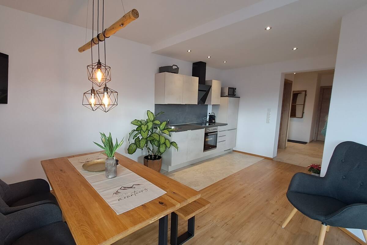 Una cocina moderna con armarios blancos, horno empotrado y microondas. Hay una mesa de comedor con dos sillas, un banco de madera y una planta en maceta. Un televisor está montado en la pared sobre la mesa.