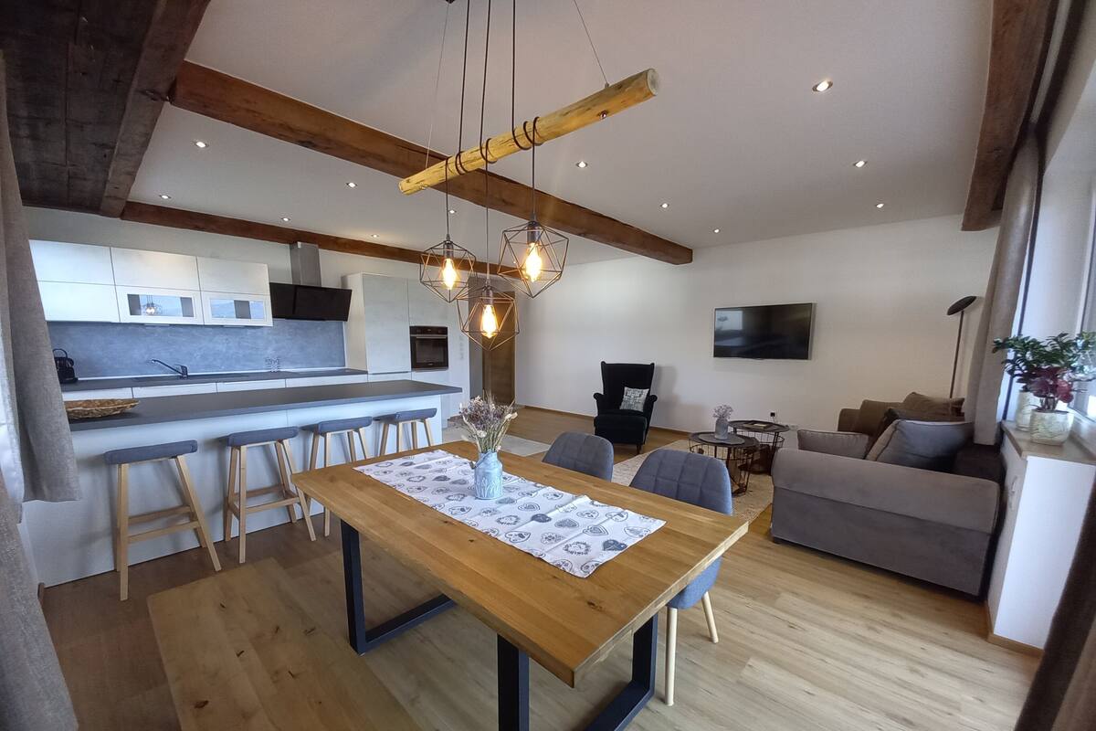 Una cocina moderna con una isla central, una zona de comedor con mesa de madera y un salón con sofá y televisor.