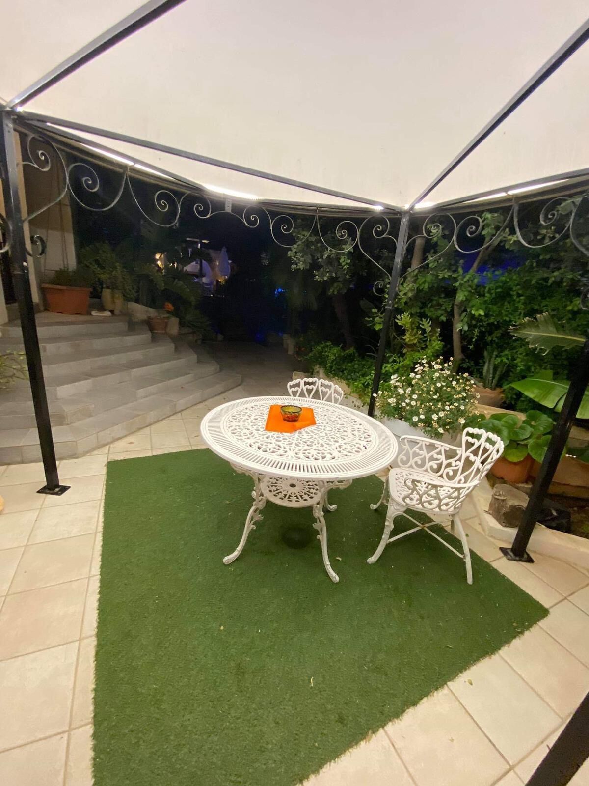 Een overdekt terras met een ronde tafel en twee stoelen, omringd door plantenpotten en een trap die naar een hoger gelegen deel leidt.