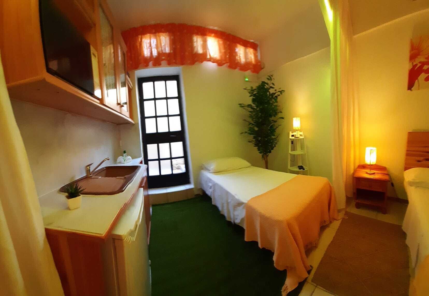 Une chambre d’hôtel compacte comprenant un petit coin cuisine, un lit simple, une table de chevet avec une lampe et une grande fenêtre donnant sur l’extérieur.