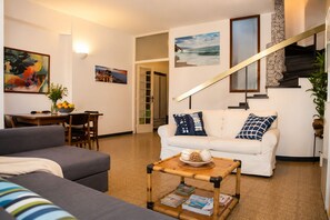 A living room with a sofa, a dining table with chairs, a staircase, and a wall with framed pictures.