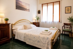Um quarto bem arrumado com cama, mesas de cabeceira, uma cadeira e uma janela com cortinas.