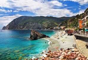 A coastal beach with clear waters, rocky outcrops, and buildings along the shore.