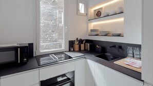 Et moderne køkken med indbygget mikroovn, ovn og vask. Et vindue med udsigt til en murstensbygning, og hylder over køkkenbordet med tallerkener og glas.