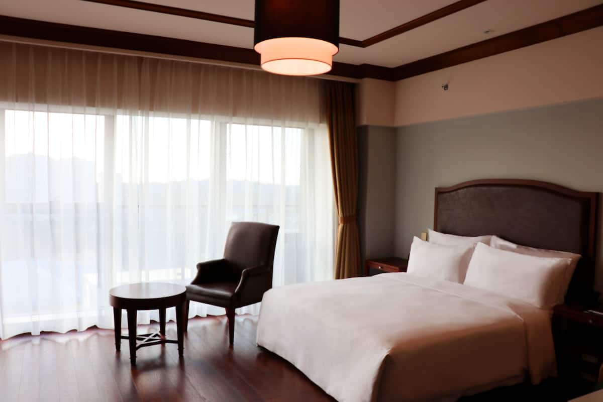 Une chambre d’hôtel avec un grand lit, une chaise, une petite table et une fenêtre avec des rideaux.
