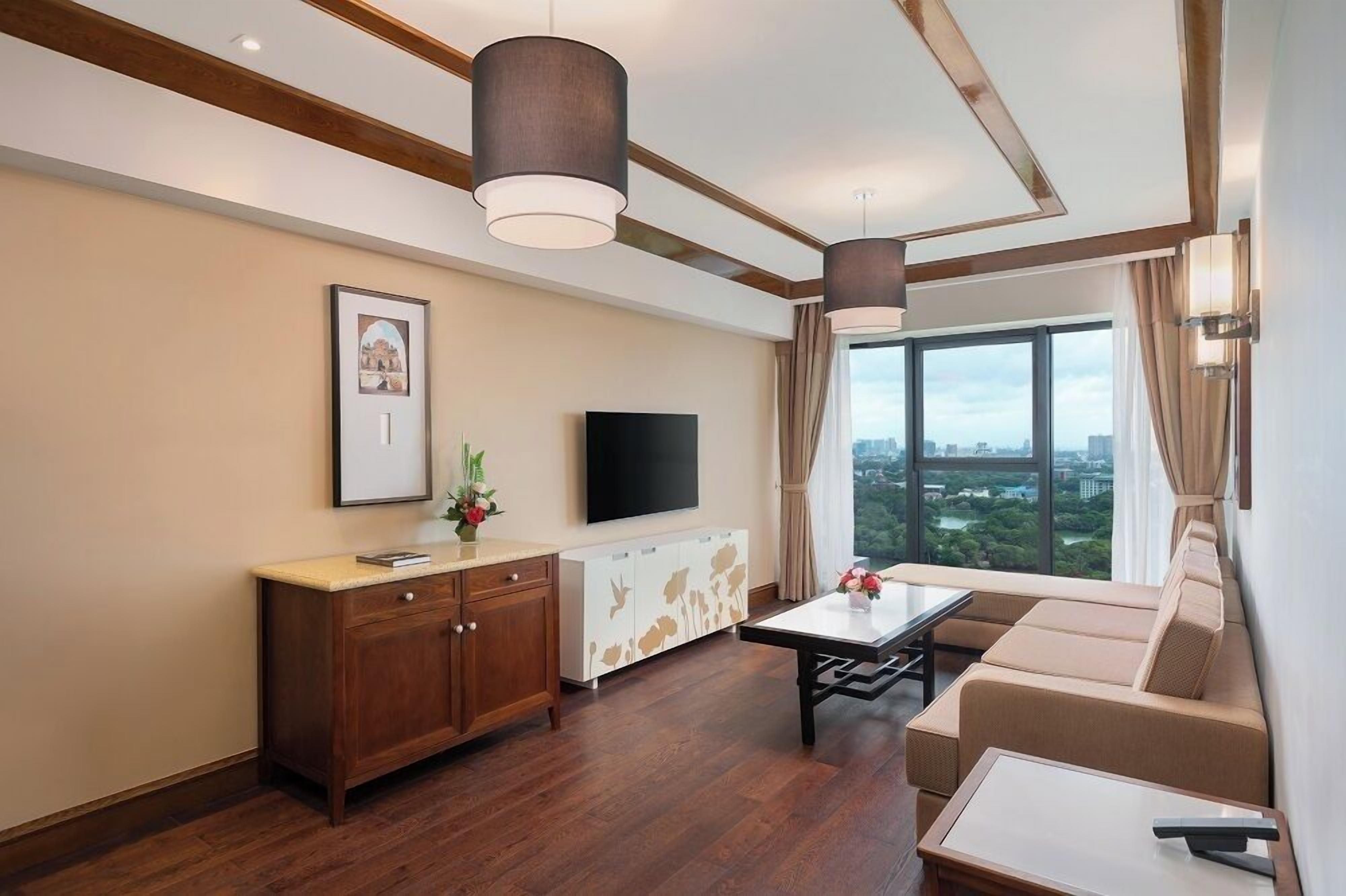 A modern hotel room with a wooden floor, a sofa, a coffee table, a TV, and a large window with a city view.